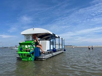 Person in a swimsuit perched on a bright green paddlewheel of a blue-and-white floating bar boat in shallow coastal water by a sandy sandbar under a sunny blue sky.