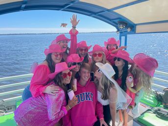 Group of friends in matching pink cowboy hats and sunglasses celebrating on a covered boat over calm blue water, holding drinks and posing close together — one person wears a white bridal veil suggesting a bachelorette boat party.
