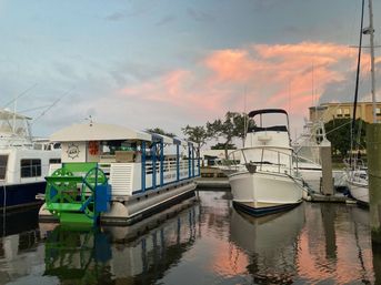 Pontoon-style floating bar with a bright green paddlewheel moored beside a white motor yacht in a calm marina, pink sunset clouds reflected on the water.