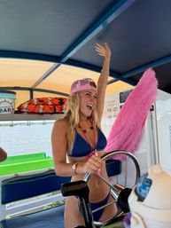 Smiling young woman in a blue bikini and pink mesh cap steering a pontoon boat with one arm raised, colorful life jackets and a fuzzy pink cushion behind her, sunny summer water and bridge in the background.