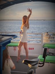 Woman in a white dress dancing barefoot on a sunset cruise boat deck, holding a canned drink with calm coastal waters and distant shoreline in the background.