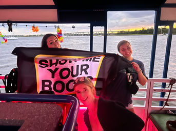 Three smiling people holding a playful banner aboard a party boat at sunset, colorful leis and pink ambient lighting, with sailboats and a bridge across the river in the background.