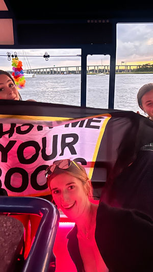 Three smiling people holding a playful banner aboard a party boat at sunset, colorful leis and pink ambient lighting, with sailboats and a bridge across the river in the background.