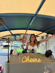 Grinning woman in a white bikini top behind a covered boat bar at a marina, holding an oversized face cutout on a stick; neon “Cheers” sign on the counter and boats visible at the dock outside.