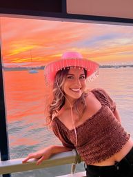 Smiling woman wearing a beaded pink cowboy hat leans on a boat railing as sailboats and a vivid orange-pink sunset reflect across a calm harbor.