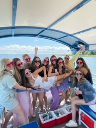 Smiling group of women in colorful sunglasses and summer outfits on a covered party boat over blue water, lifting a friend in a white dress above coolers with drinks on a sunny day