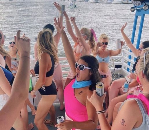 Group of women partying on a pontoon boat on a sunny lake, wearing swimsuits and sunglasses, holding canned drinks and cheering with arms raised