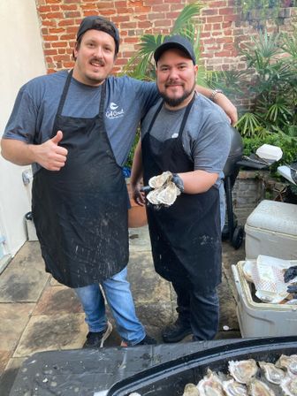 Two apron-clad men smiling on a brick-walled urban patio holding freshly shucked oysters over a tray, thumbs-up beside a grill and coolers — backyard oyster shucking scene.
