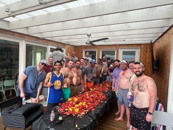 Group of friends at a backyard crawfish boil on a covered wooden deck, gathered around a long table piled with red crawfish, corn, potatoes and drinks for a casual summer seafood party.