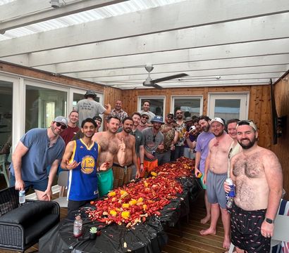 Group of friends at a backyard crawfish boil on a covered wooden deck, gathered around a long table piled with red crawfish, corn, potatoes and drinks for a casual summer seafood party.