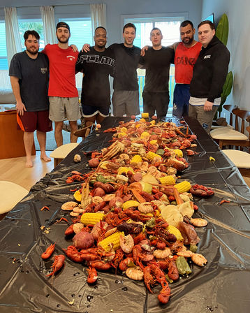 Group of seven people standing behind a long indoor table piled with a Cajun-style seafood boil — crawfish, shrimp, crab legs, corn on the cob, red potatoes and sausage on a black tablecover.