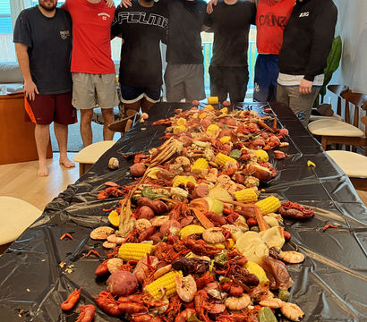 Group of seven people standing behind a long indoor table piled with a Cajun-style seafood boil — crawfish, shrimp, crab legs, corn on the cob, red potatoes and sausage on a black tablecover.
