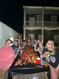 Group of friends on a wooden deck at night eating a lively crawfish boil piled with crawfish, corn, and potatoes at a long table in front of a two-story house with balcony.