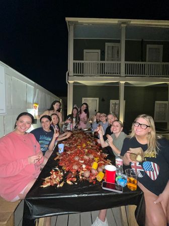 Group of friends on a wooden deck at night eating a lively crawfish boil piled with crawfish, corn, and potatoes at a long table in front of a two-story house with balcony.