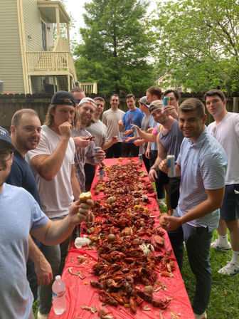 Group of friends enjoying a backyard crawfish boil in a suburban yard, long red-covered table piled high with boiled crawfish, corn and potatoes as people smile and hold drinks.