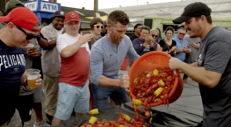 Man pouring a large orange basket of cooked crawfish, corn and potatoes onto a table at an outdoor seafood boil festival while a cheering crowd watches with drinks.