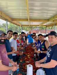 Smiling group at a lively outdoor crawfish boil under a metal‑roof park pavilion, long picnic table piled with red crawfish, corn, potatoes and drinks.