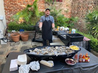 Outdoor patio oyster station in a brick courtyard: server stands behind a boat-shaped tray of ice filled with raw oysters, lemon wedges, sauces and disposable plates and forks.