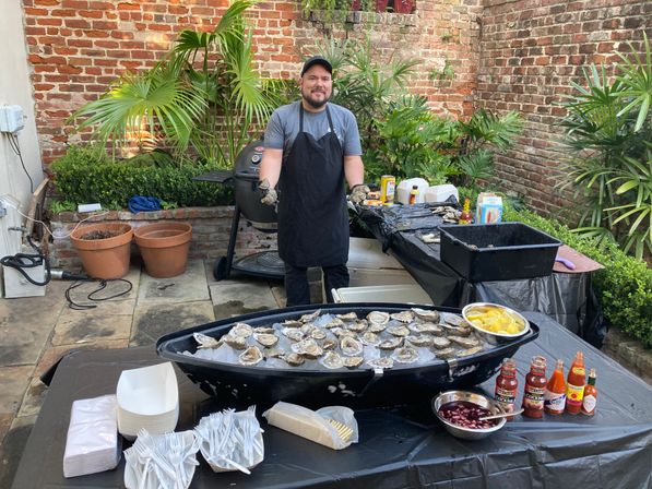 Outdoor patio oyster station in a brick courtyard: server stands behind a boat-shaped tray of ice filled with raw oysters, lemon wedges, sauces and disposable plates and forks.