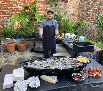 Outdoor patio oyster station in a brick courtyard: server stands behind a boat-shaped tray of ice filled with raw oysters, lemon wedges, sauces and disposable plates and forks.