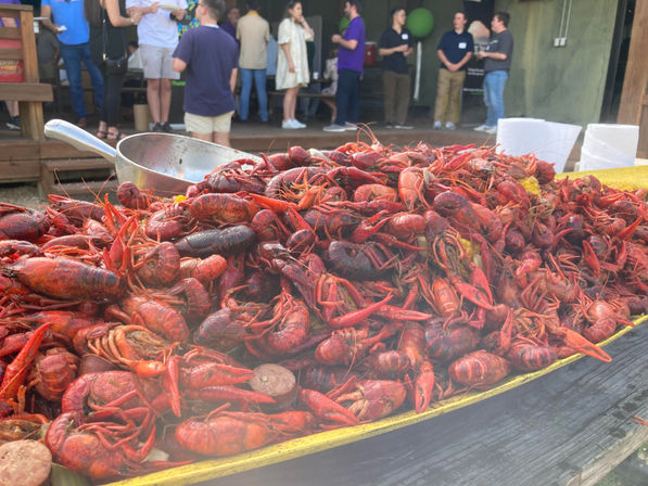 Cajun-style pile of bright red boiled crawfish with corn and sausage on a yellow tray, metal scoop and paper towels at an outdoor social gathering