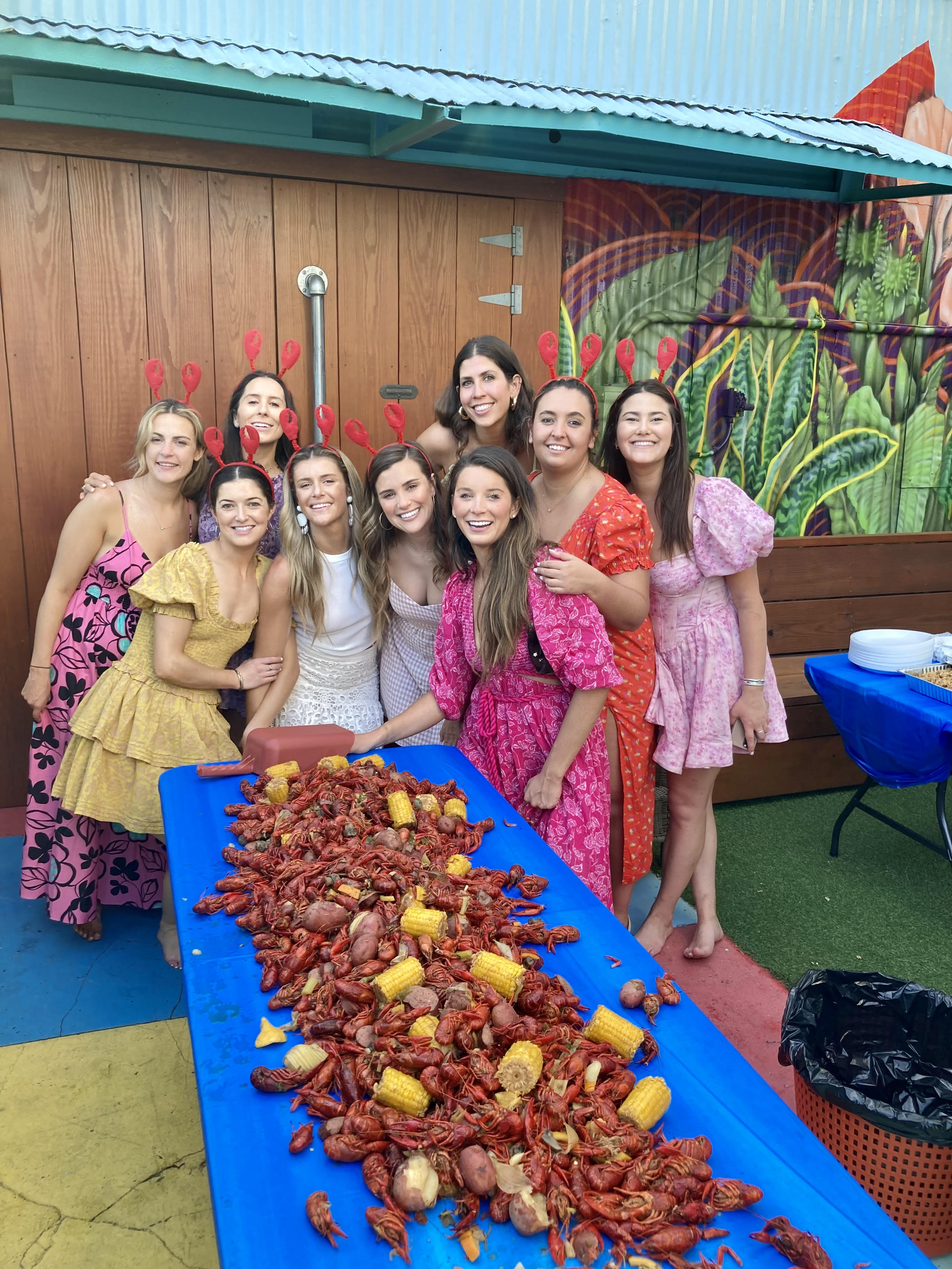 Group of women in colorful dresses and red crawfish headbands smiling around a long blue table piled with crawfish, corn and potatoes at a festive outdoor crawfish boil on a muraled patio.