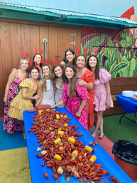 Group of women in colorful dresses and red crawfish headbands smiling around a long blue table piled with crawfish, corn and potatoes at a festive outdoor crawfish boil on a muraled patio.