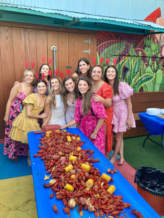 Group of women in colorful dresses and red crawfish headbands smiling around a long blue table piled with crawfish, corn and potatoes at a festive outdoor crawfish boil on a muraled patio.
