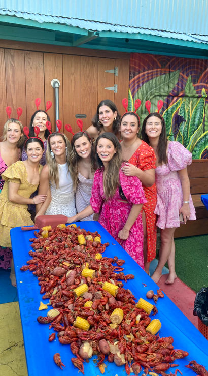 Group of women in colorful dresses and red crawfish headbands smiling around a long blue table piled with crawfish, corn and potatoes at a festive outdoor crawfish boil on a muraled patio.