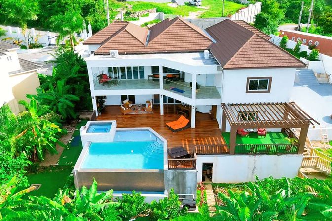 Aerial view of a modern two-story tropical villa on a green hillside with brown-tiled roofs, a wooden deck with orange loungers, a pergola seating area, and an elevated infinity pool and spa surrounded by lush banana and palm foliage.