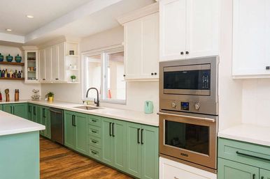 Bright modern kitchen with sage-green lower cabinets, white upper cabinets, white countertops, stainless steel built-in microwave and oven, farmhouse sink under a window, wood floors and open shelving with decor.