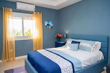 Sunlit coastal-style bedroom with blue walls and upholstered queen bed, yellow curtains, wall air conditioner, nightstand lamp and fresh flowers
