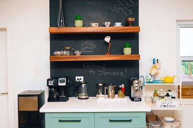 Cheerful coffee station with mint-green cabinets, white countertop, two coffee makers and kettle, wooden floating shelves with mugs and potted plants, and a black chalkboard that reads 'Welcome to Jamaica!'.