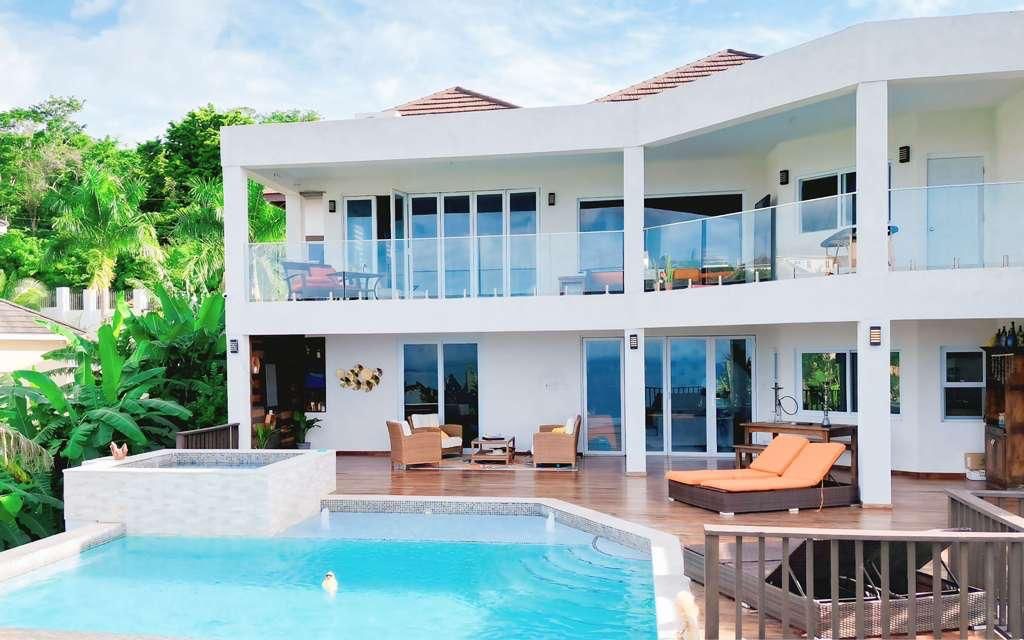 Modern two-story tropical villa with glass balconies, wooden deck, orange lounge chairs and a clear blue pool with lush greenery and ocean views.
