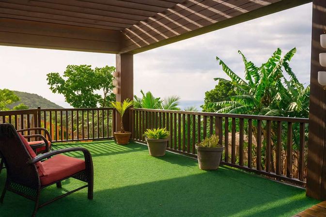 Shaded tropical deck with green turf, wicker lounge chairs with red cushions and potted palms, wooden railing framing banana trees, hillside and a distant ocean view.