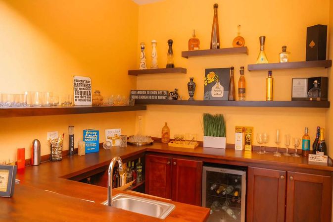 Cozy home bar nook with warm yellow walls, wooden countertop and corner sink, dark floating shelves holding liquor bottles, glassware, decorative signs, a small wine fridge and a potted grass planter.