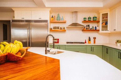 Bright modern kitchen with green lower cabinets, stainless double-door refrigerator, stainless range hood, white quartz island with wood breakfast bar and gooseneck faucet, open wood shelves, and a fruit bowl of bananas.