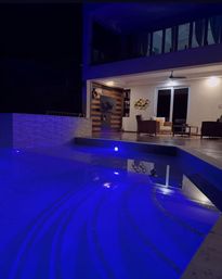 Modern backyard patio with wicker lounge chairs and sliding glass doors beside a curved swimming pool glowing electric blue at night, lit by ambient terrace lights.