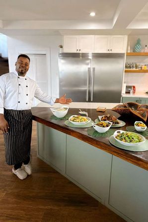 Chef in white jacket and striped apron gestures toward colorful salads and roasted dishes displayed on a wooden-topped island in a modern home kitchen with stainless steel refrigerator.