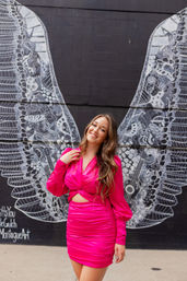 Young woman in a bright fuchsia ruched dress smiling and posing in front of a large white angel-wings street-art mural on a black brick wall, playful urban portrait.