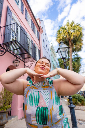 Playful woman in a colorful patterned dress posing with hands under her chin on a sunny, palm-lined sidewalk in front of pastel row houses, wrought-iron balconies, and a vintage streetlamp.