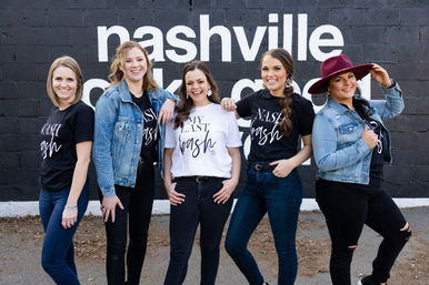 Five smiling women in coordinated 'Nashville' and 'My Last Bash' shirts and denim jackets posing in front of a black brick wall painted with 'Nashville' — playful Nashville bachelorette group photo.