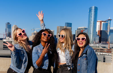 Four friends in denim jackets wearing heart-shaped sunglasses laugh and pose on a sunny rooftop with a modern city skyline and blue sky in the background.