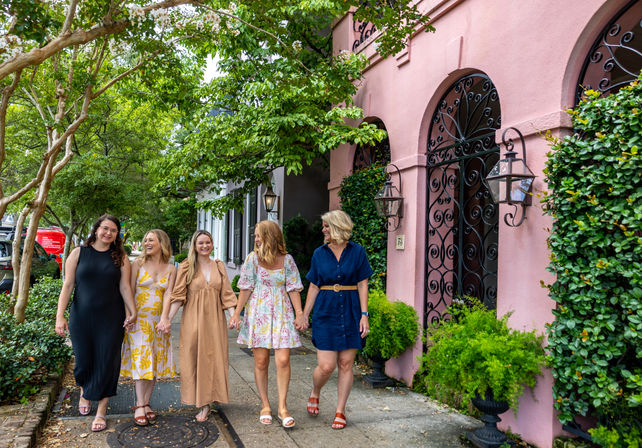 Five women friends strolling and laughing along a tree-lined sidewalk past a pink stucco building with arched wrought-iron gates and lanterns.