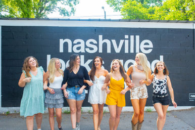Seven friends laughing arm-in-arm walking in front of a large “Nashville” mural, wearing bright summer dresses, shorts and boots on a sunny day