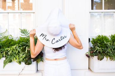 Back-facing woman in white outfit holding a wide-brim sun hat that reads "Mrs. Carter", standing in front of white windows with lush green window-box plants.
