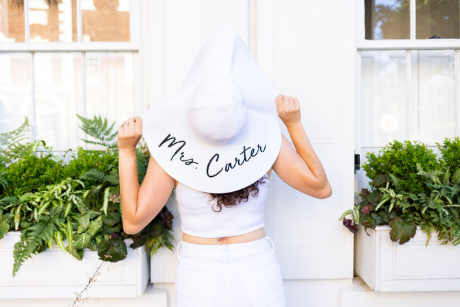 Back-facing woman in white outfit holding a wide-brim sun hat that reads "Mrs. Carter", standing in front of white windows with lush green window-box plants.