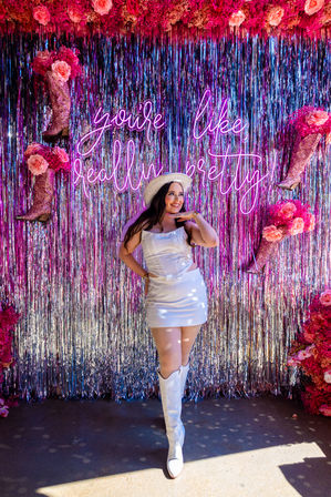 Smiling person in a white mini dress, cowboy hat and white boots posing at a pink metallic fringe event photo booth with neon sign 'you're like, really pretty', glittery pink boot props and roses.