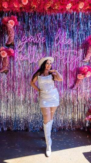 Smiling person in a white mini dress, cowboy hat and white boots posing at a pink metallic fringe event photo booth with neon sign 'you're like, really pretty', glittery pink boot props and roses.