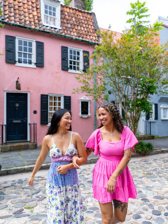 Two smiling women arm-in-arm strolling a cobblestone street past pink pastel row houses and greenery, one in a floral maxi and the other in a bright pink dress.